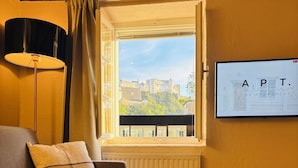 A cozy room with a sofa, a round table, a window with a view of a castle, and a wall-mounted sign.