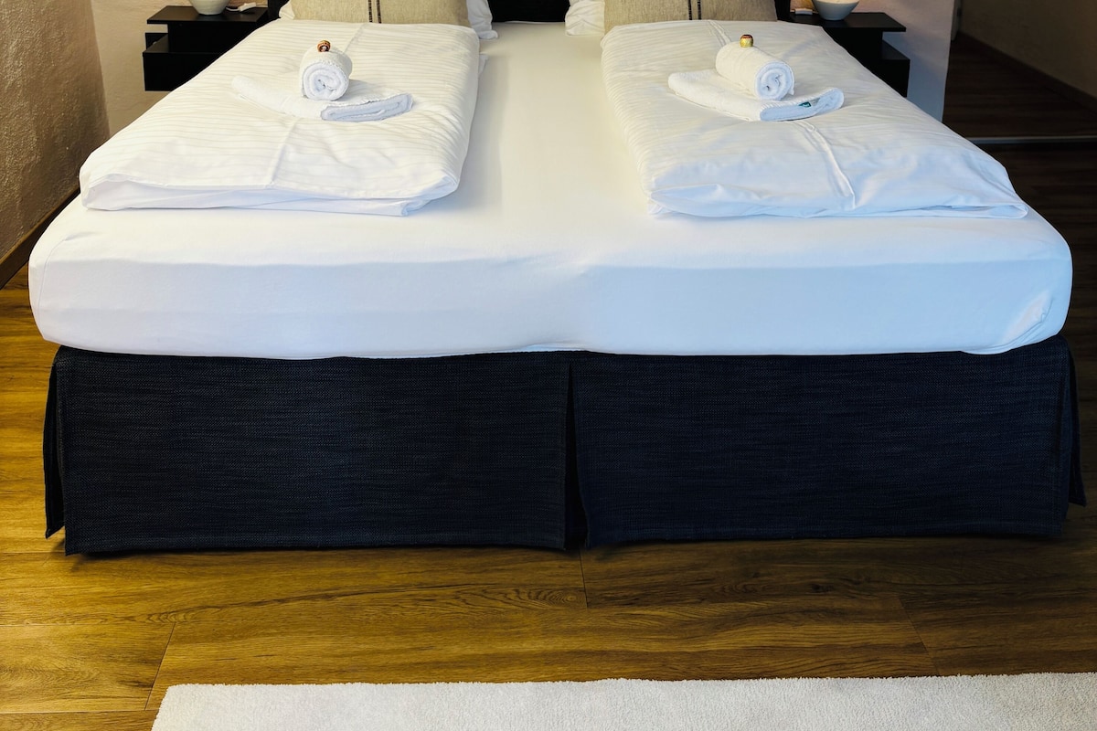 A double bed with white linens, two nightstands with lamps, and two wall-mounted paintings.