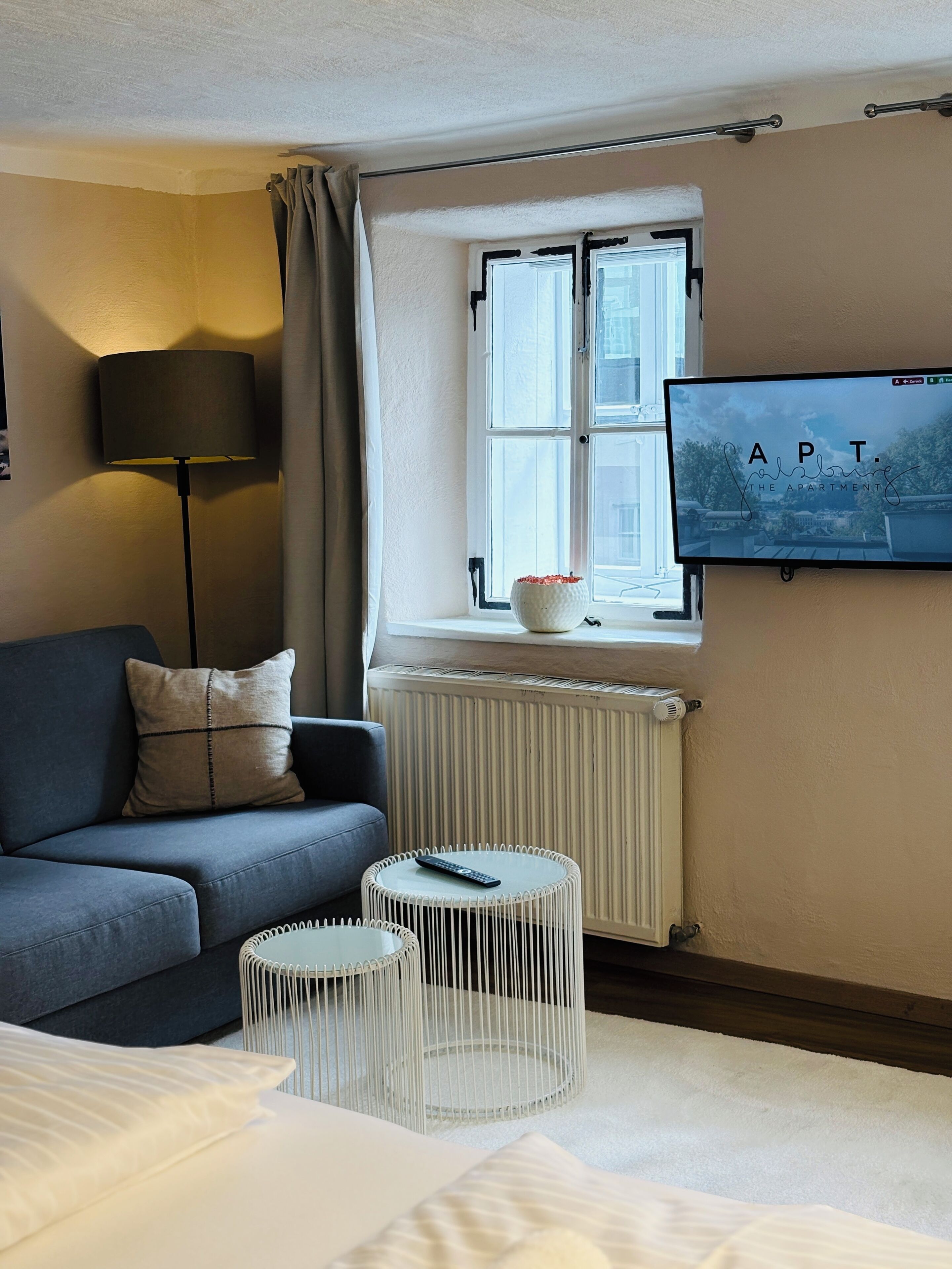 A modern hotel room with a grey sofa, a small round table, a bed, a radiator, and a TV mounted on the wall.