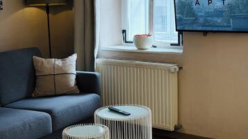 A modern hotel room with a grey sofa, a small round table, a bed, a radiator, and a TV mounted on the wall.