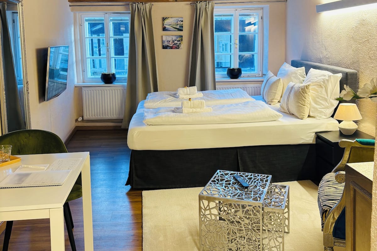 A bedroom with a wooden ceiling, a bed with white linens, a bedside table, a chair, and a window with curtains.