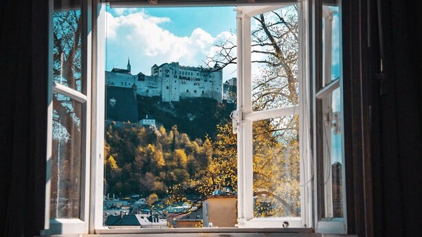 A view through a window of a castle on a hill, with autumn trees in the foreground.
