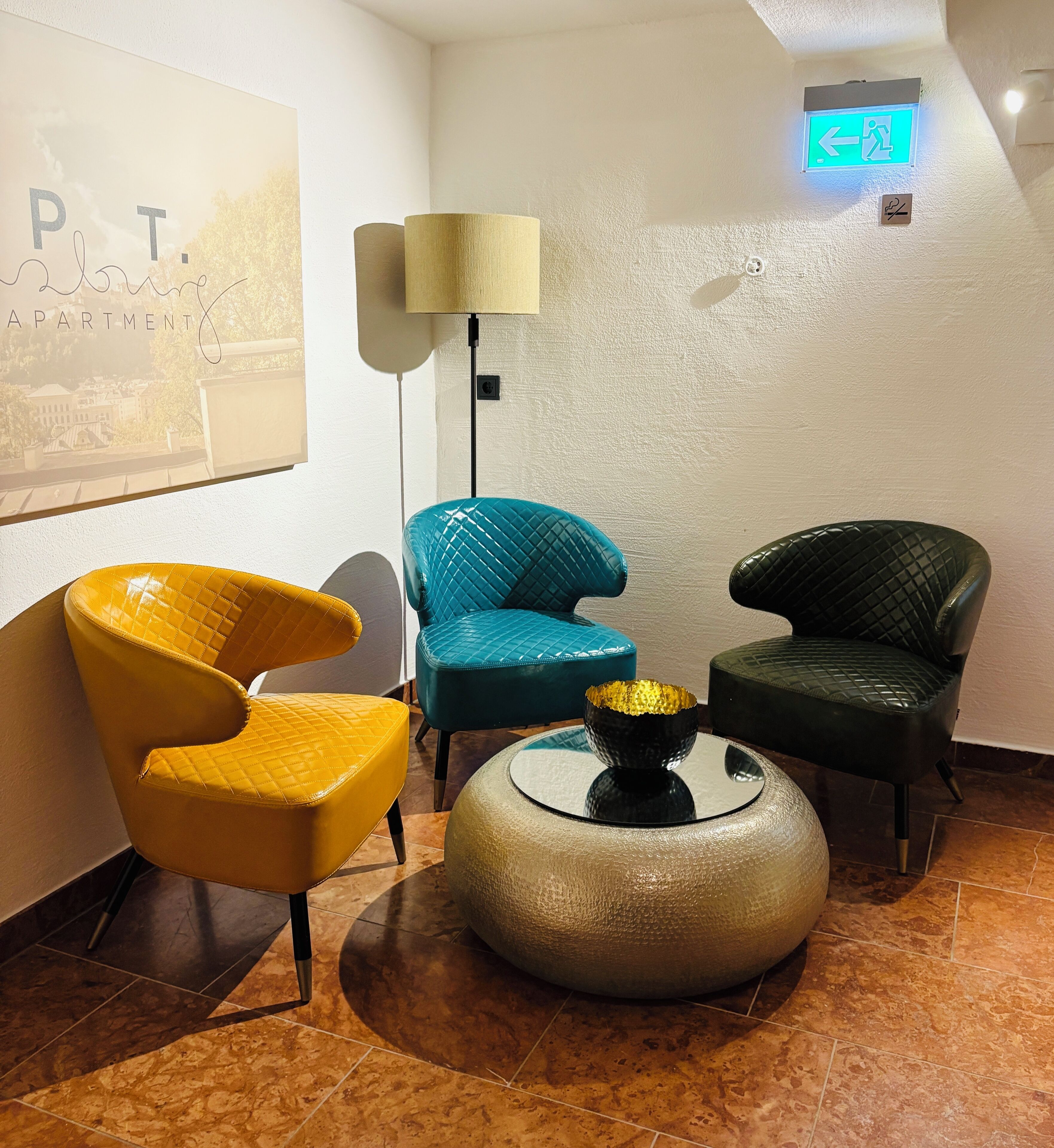 Un hall d’entrée moderne avec un fauteuil jaune, un fauteuil turquoise et un fauteuil noir. On y trouve une table ronde sur laquelle est posé un vase décoratif. Une lampe sur pied et un panneau indiquant la sortie sont également visibles.