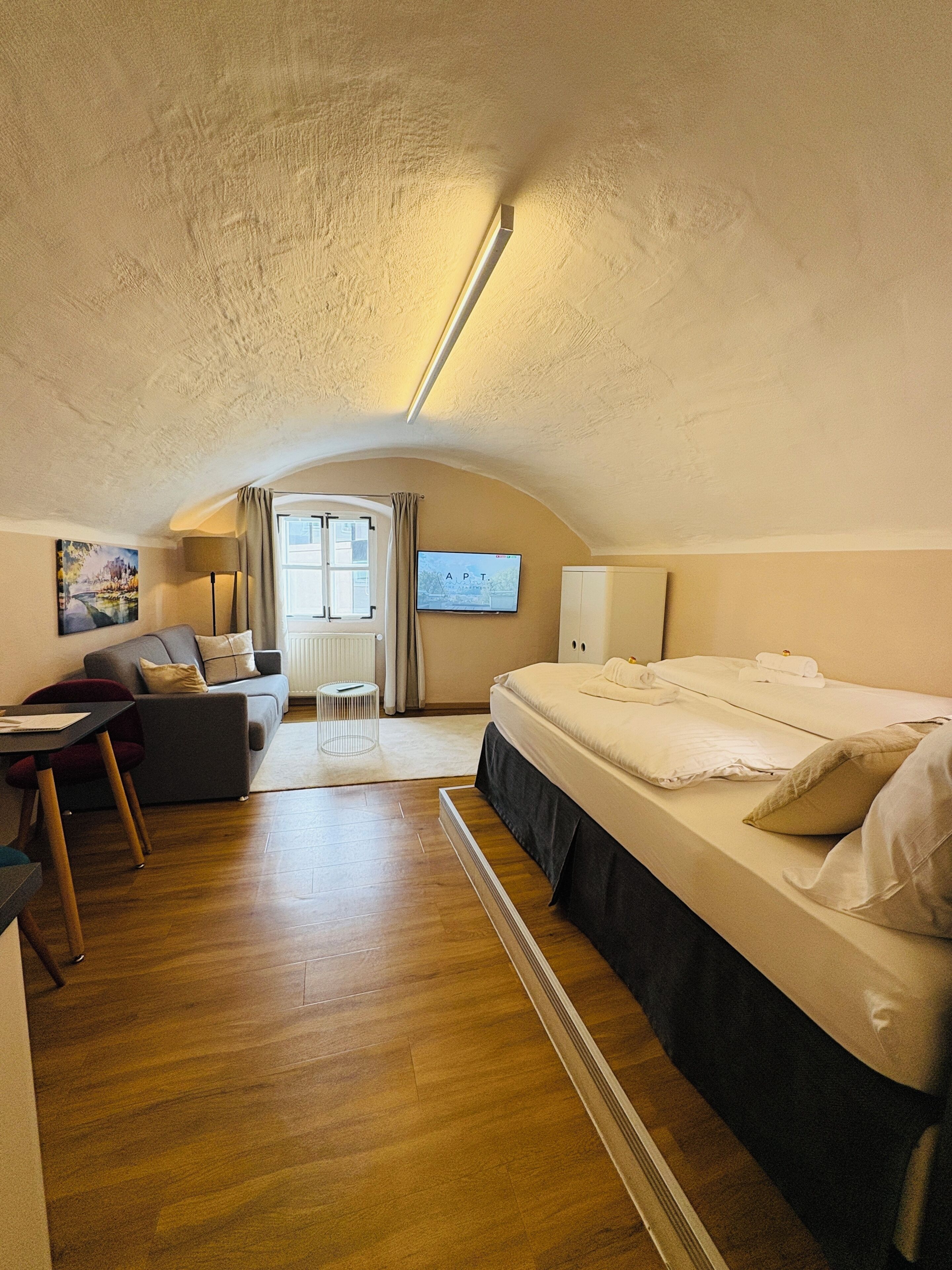 A cozy, well-lit room with a bed, a sofa, a small table, and a chair. There is a window with curtains, a painting on the wall, and a flat-screen TV.