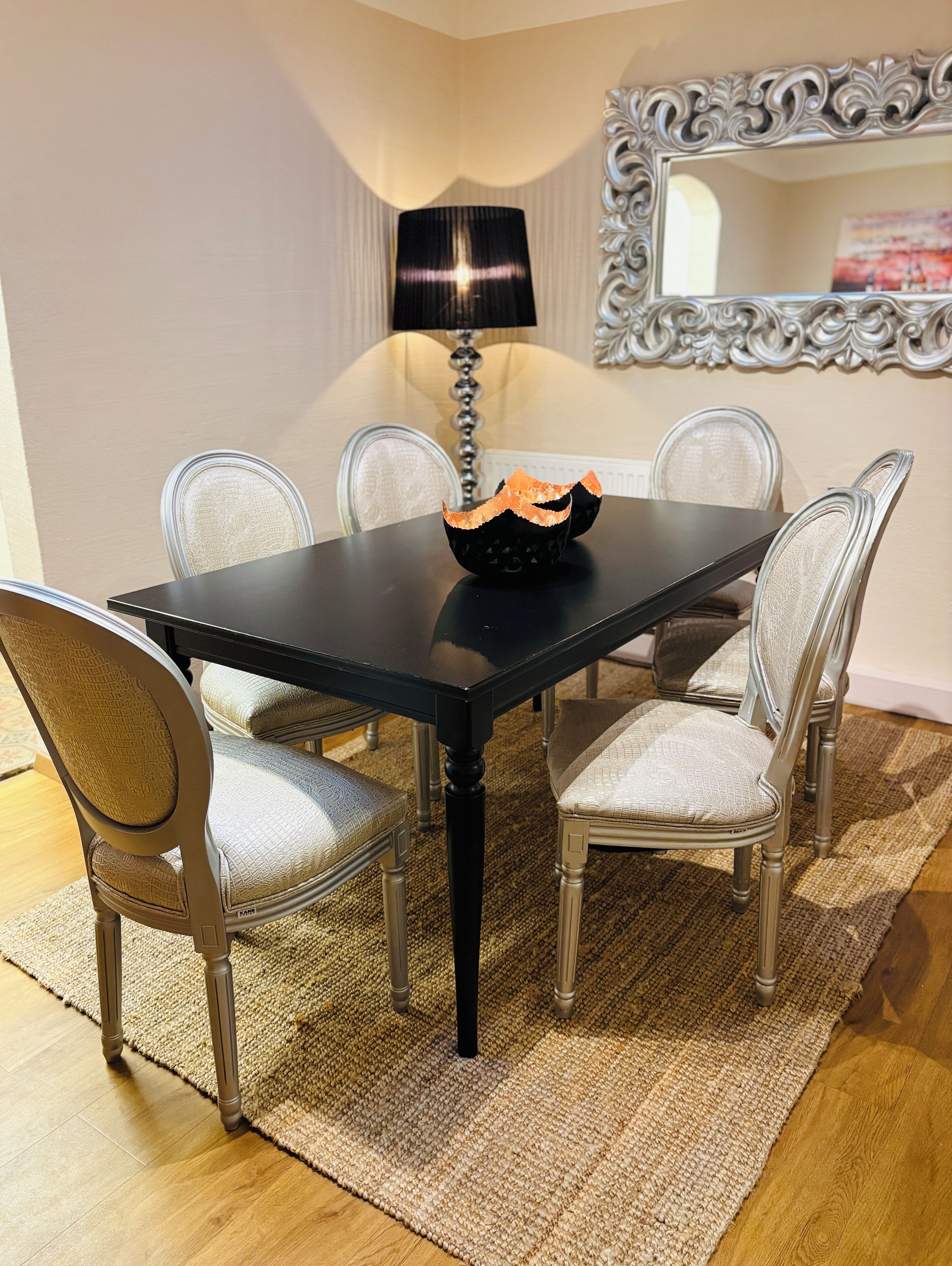 A dining area with a black table, six chairs, a lamp, and a decorative mirror.