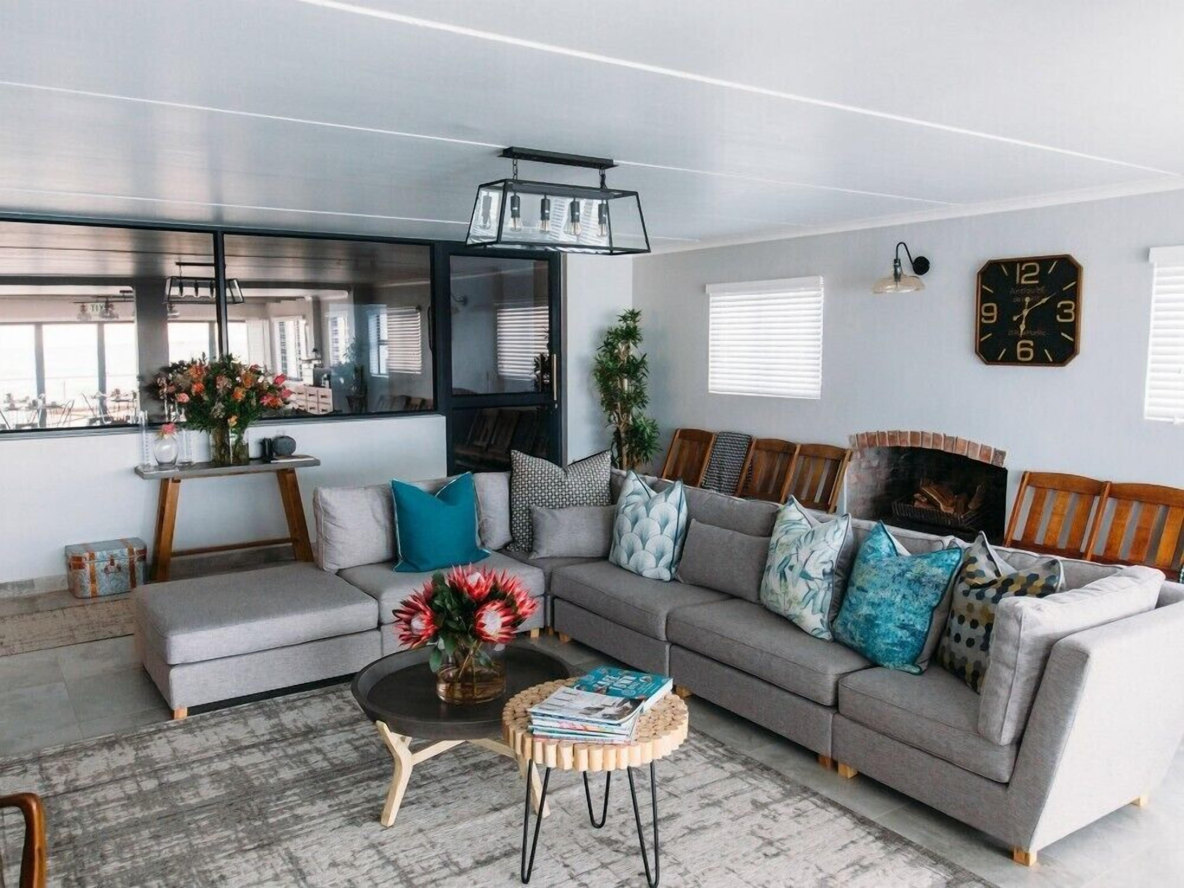 A modern living room with a grey sofa, a coffee table, and a bookshelf. There is a window with blinds, a clock on the wall, and a plant on the floor.
