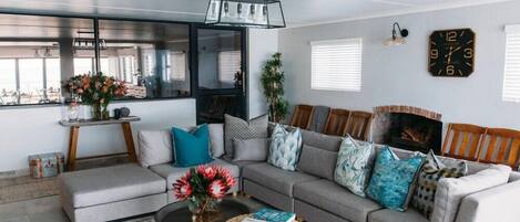A modern living room with a grey sofa, a coffee table, and a bookshelf. There is a window with blinds, a clock on the wall, and a plant on the floor.