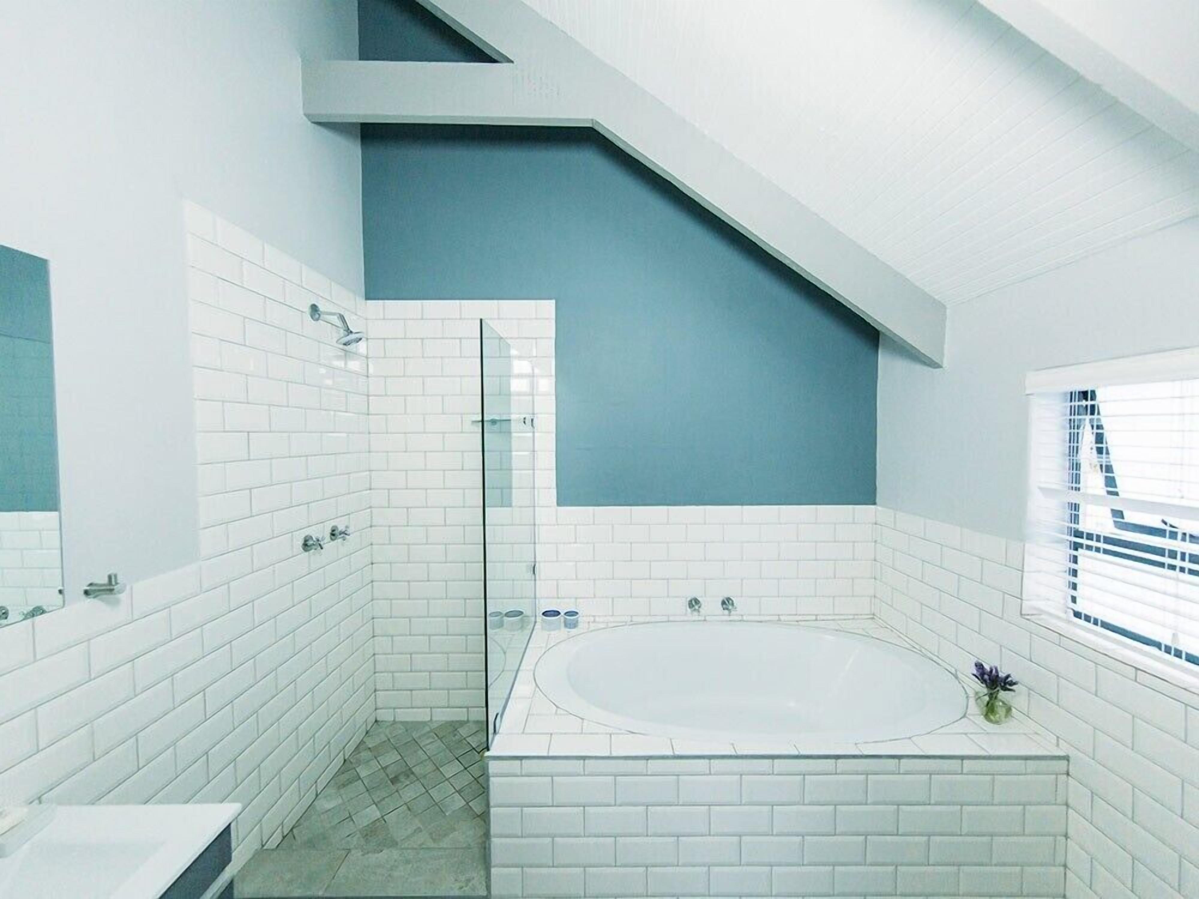 Une salle de bain moderne avec des carreaux blancs, une douche en verre, une baignoire sur pieds et une fenêtre avec des stores.