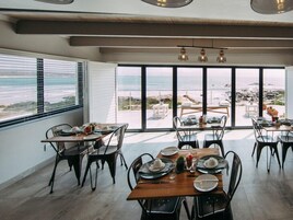 Un comedor con mesas dispuestas para cuatro personas, ventanas amplias que ofrecen vista a la playa y una decoración de diseño moderno.