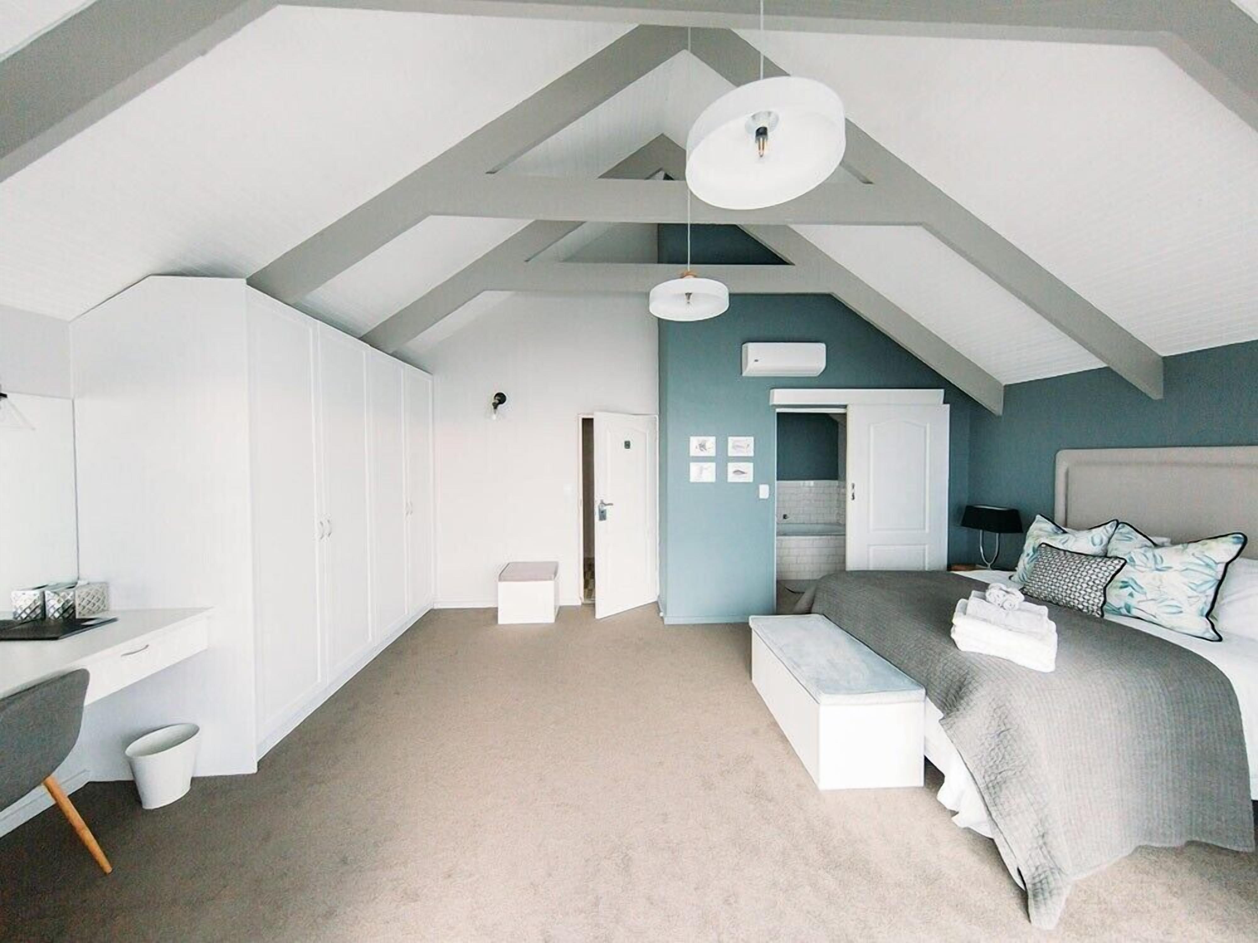 A modern bedroom with a sloped ceiling, a bed with a grey headboard, a desk with a chair, and a large wardrobe.