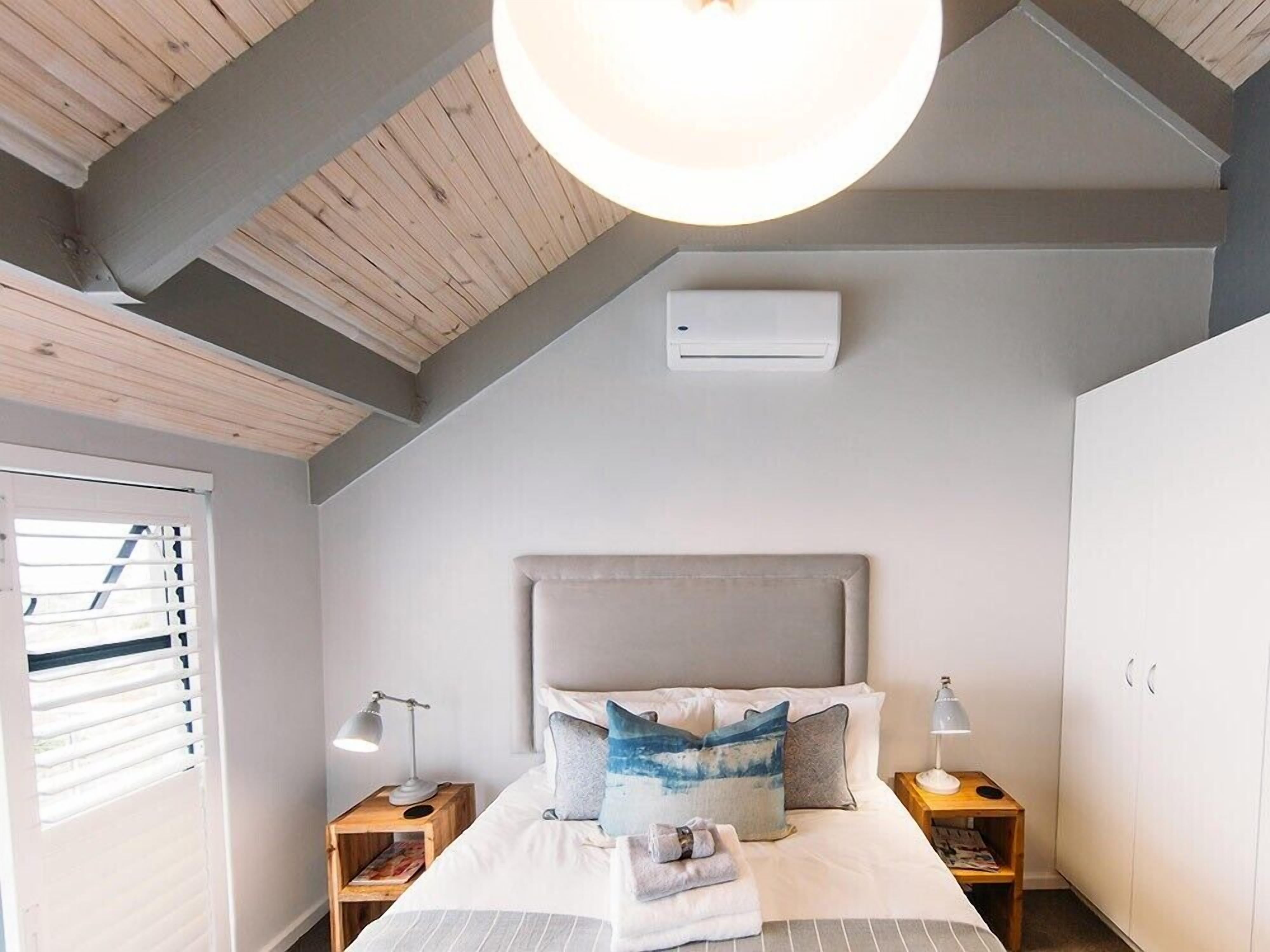 A bedroom with a sloped ceiling, a bed with pillows, bedside lamps, a wardrobe, and an air conditioning unit.