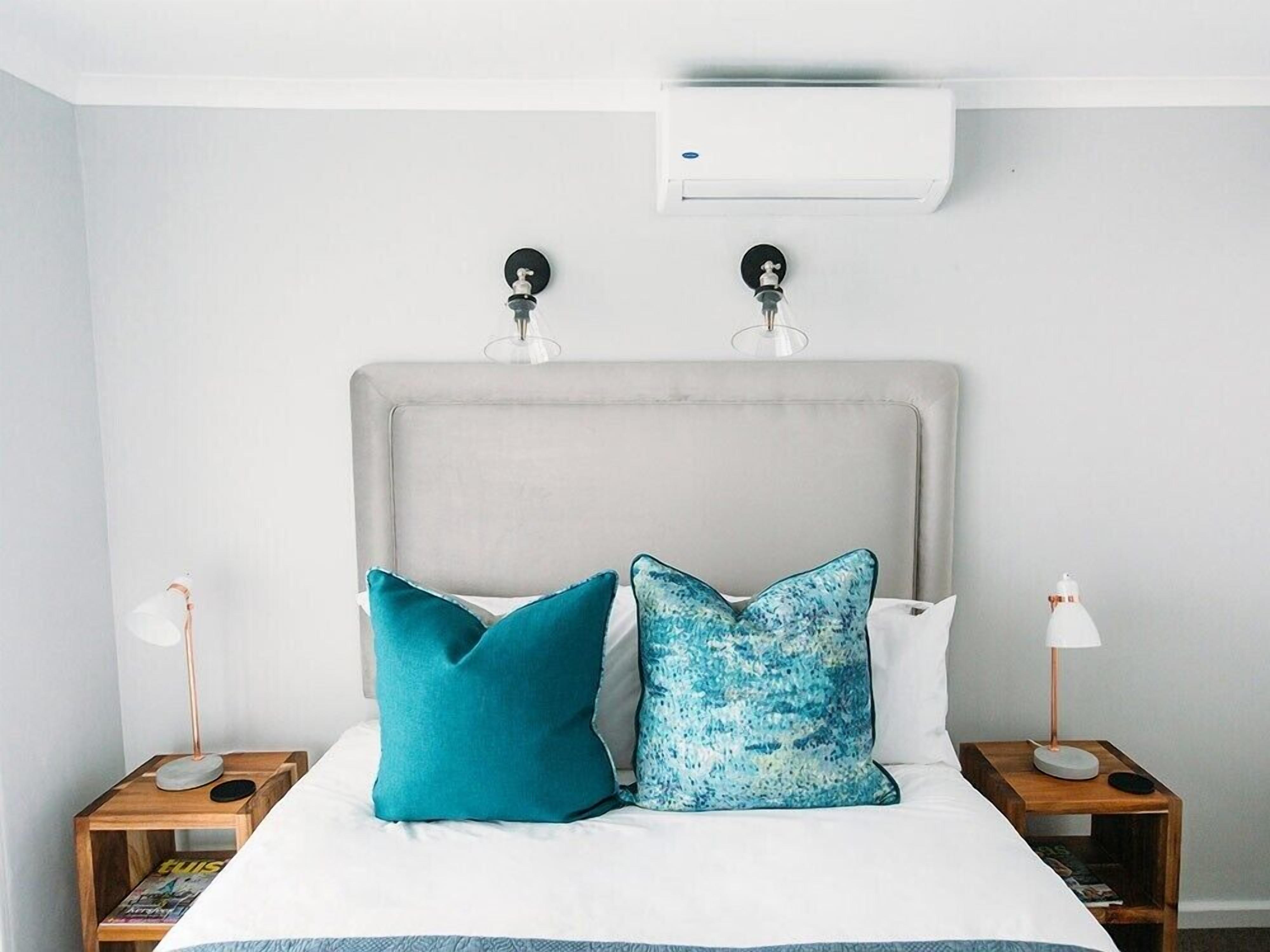 A neatly made bed with two pillows, a grey upholstered headboard, and an air conditioning unit above.
