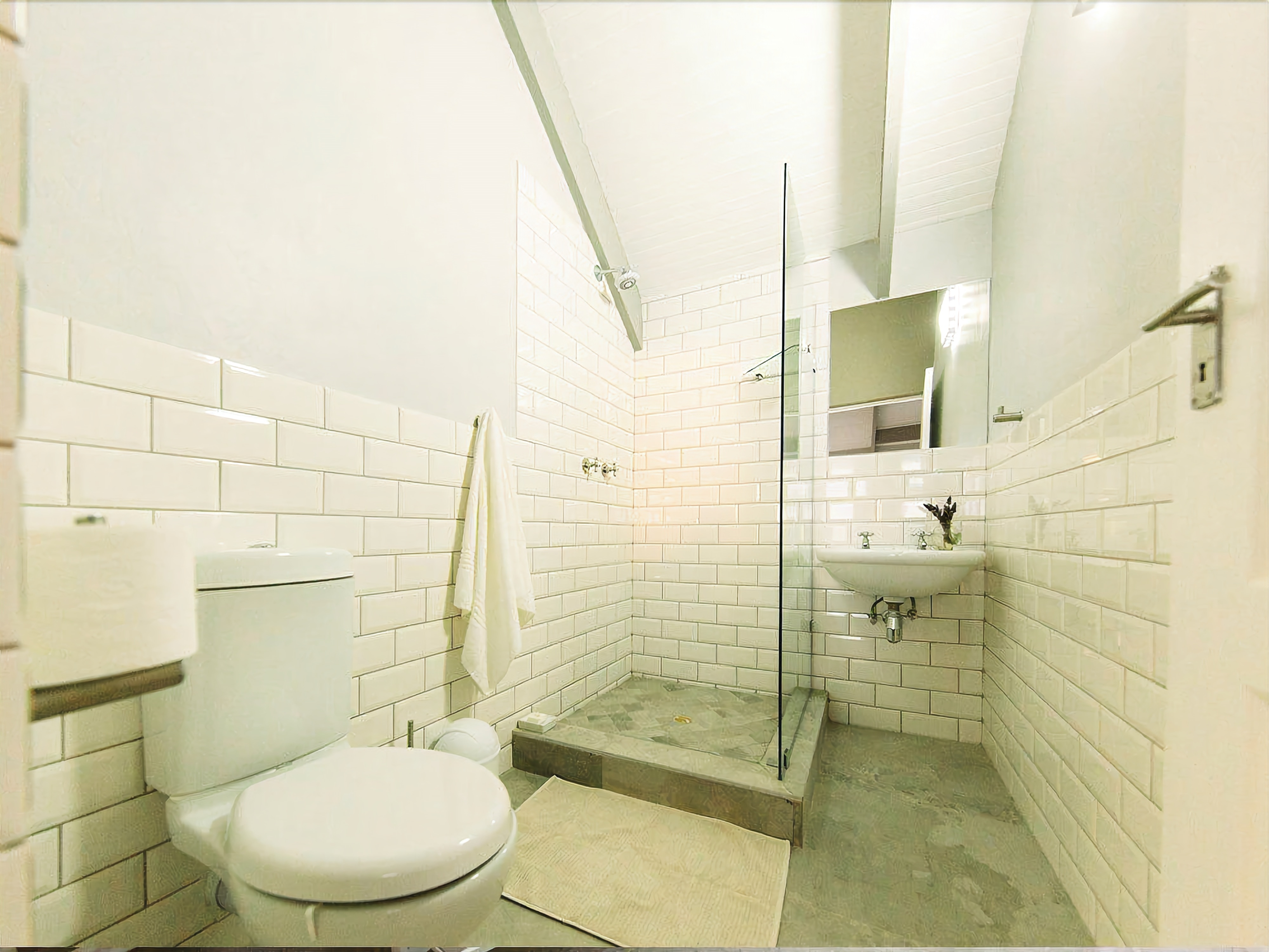 Un baño pequeño con azulejos blancos, inodoro, cabina de ducha con mampara de vidrio y lavamanos.