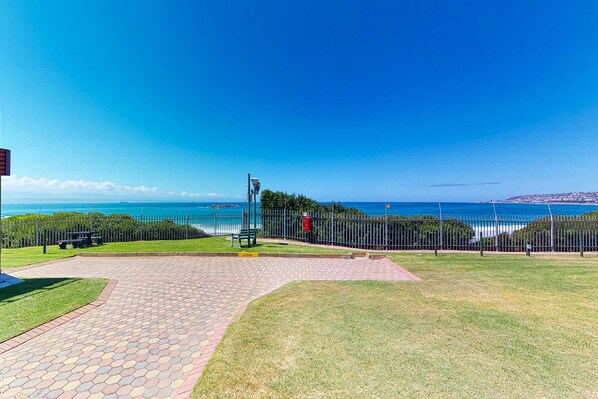 A paved pathway leading to a grassy area with a metal fence and a bench, overlooking a coastal view.