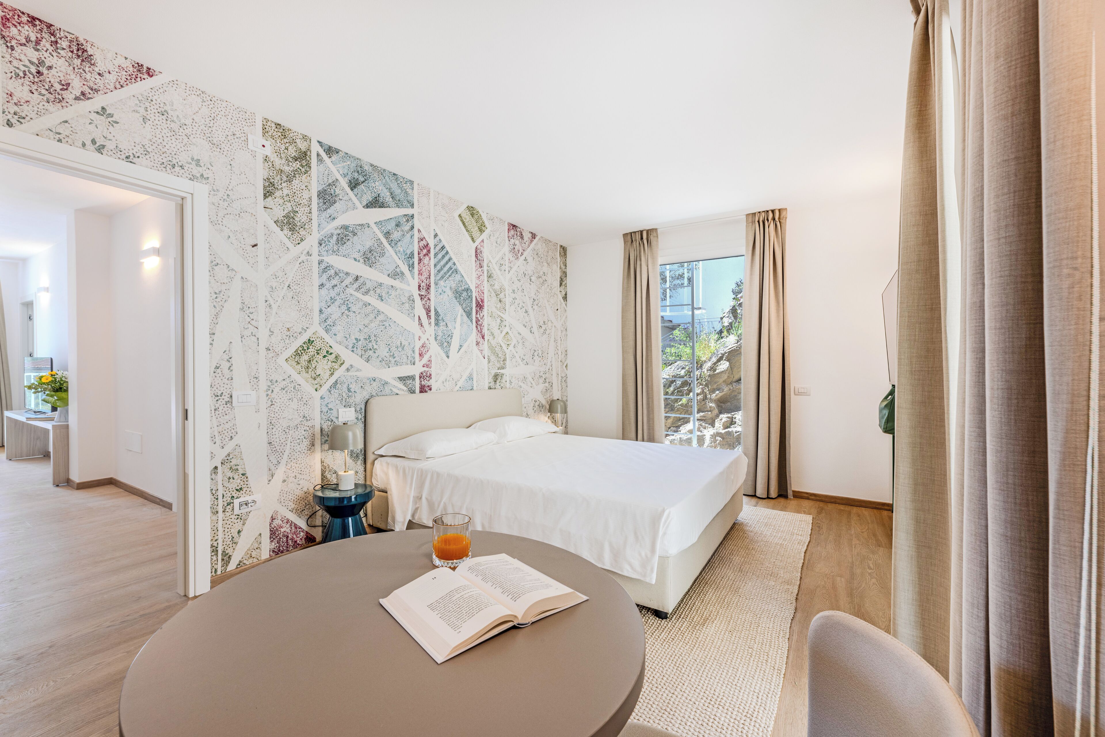 Ein Hotelzimmer mit einem Bett, einem runden Tisch mit einem Buch und einem Glas Orangensaft, sowie Blick ins Freie durch das Fenster.