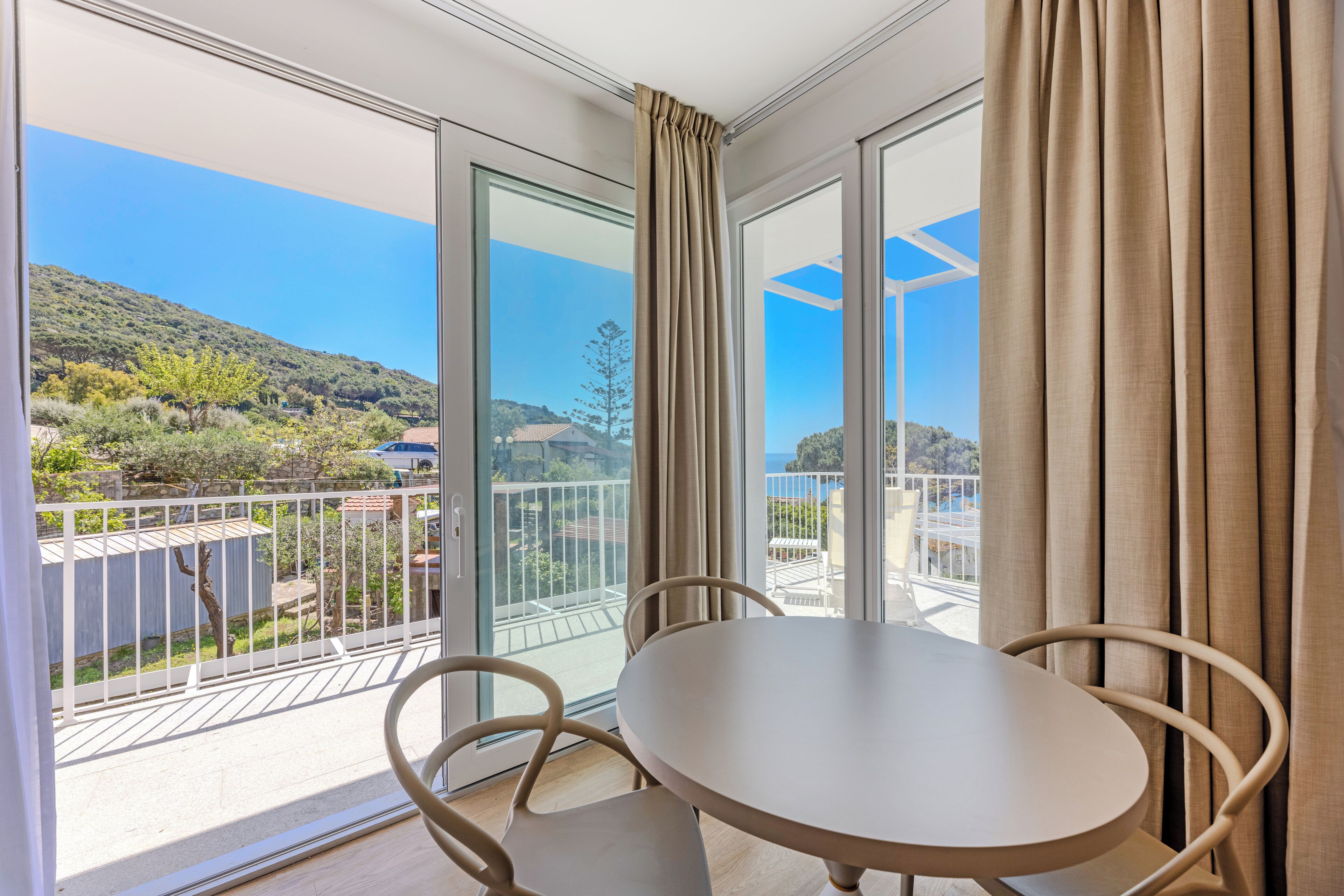 A balcony with a table and chairs, offering a view of a hillside and a distant coastline.