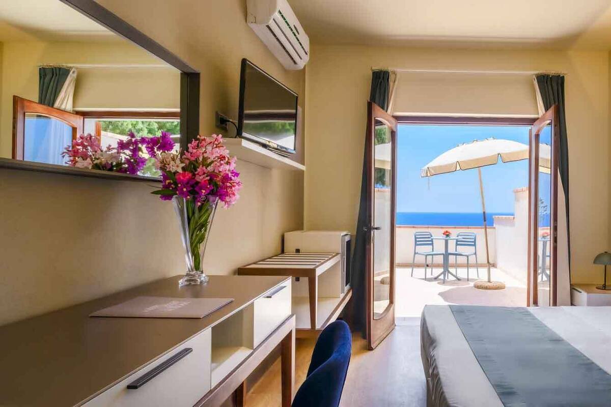 Een hotelkamer met een bed, een bureau, een televisie, een bloevas en een balkon met uitzicht op zee.