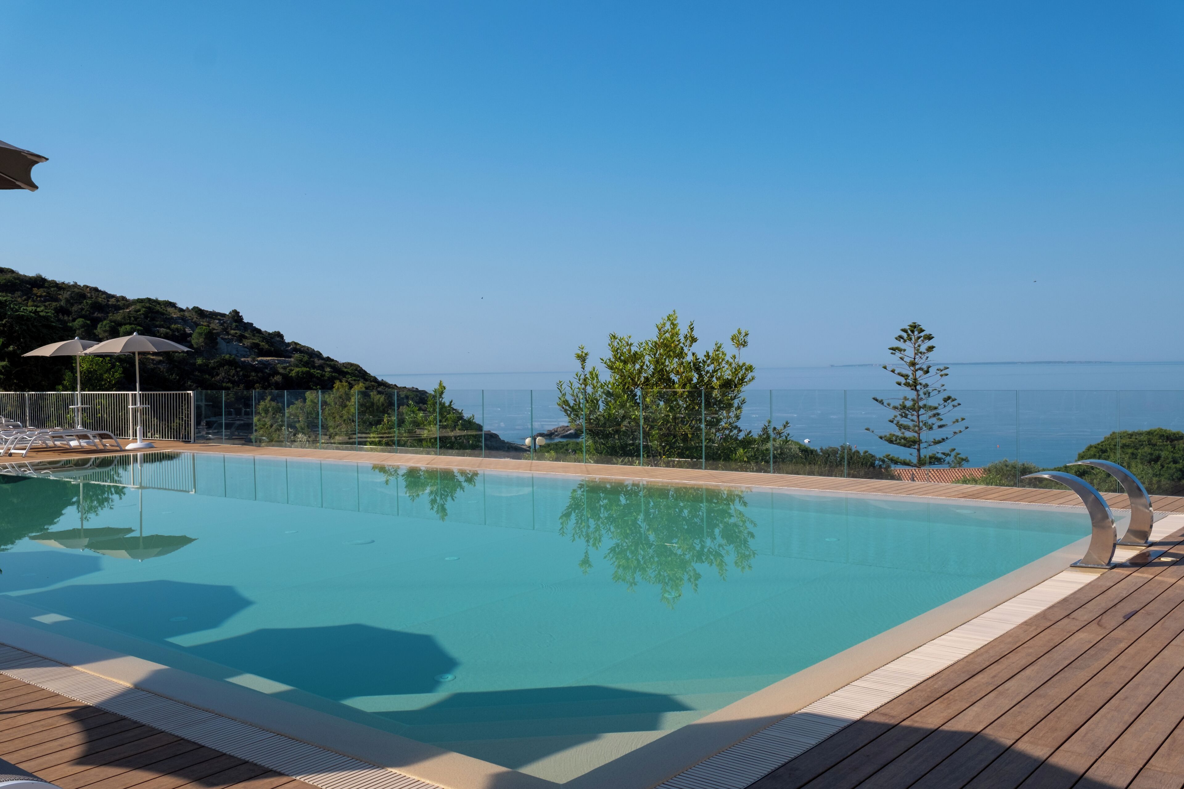 A clear swimming pool with a wooden deck, lounge chairs, and a view of the sea.