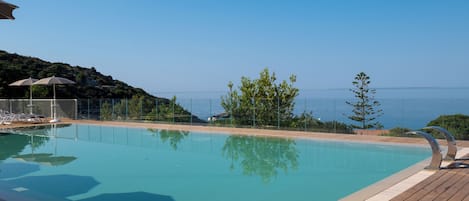 A clear swimming pool with a wooden deck, lounge chairs, and a view of the sea.