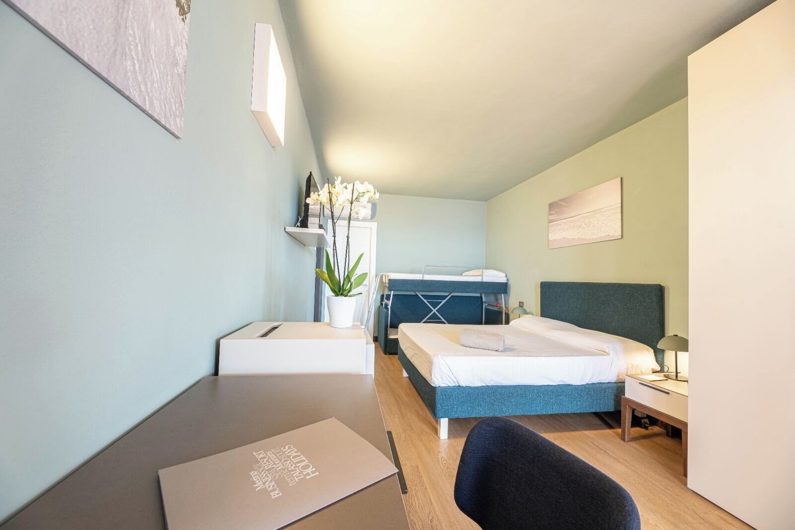 A compact hotel room with a bunk bed, a desk with a laptop, a chair, a bedside table with a lamp, and a potted plant.