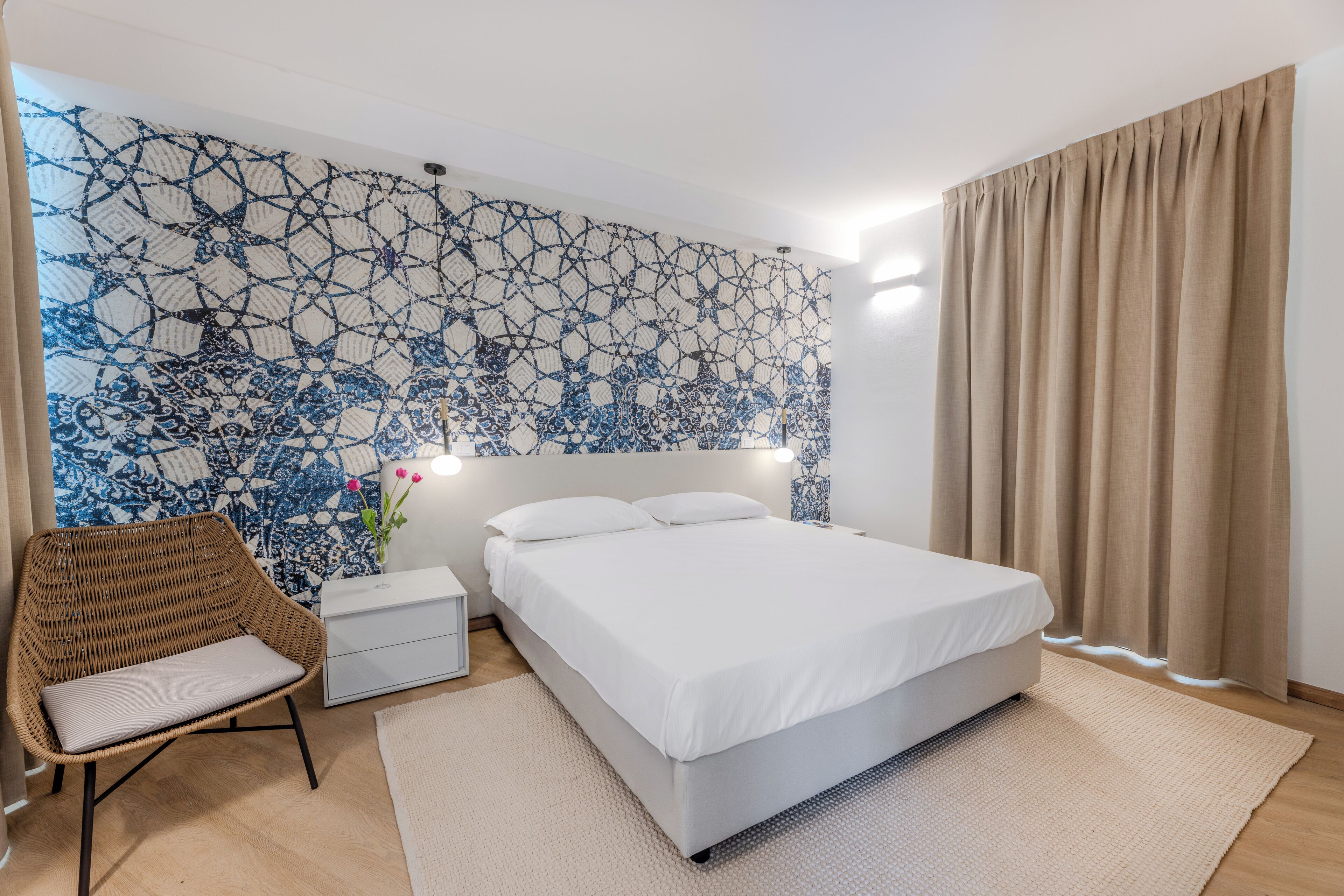 A bedroom with a bed, a nightstand, a wicker chair, a white bedside table, a patterned wall, and a window with beige curtains.