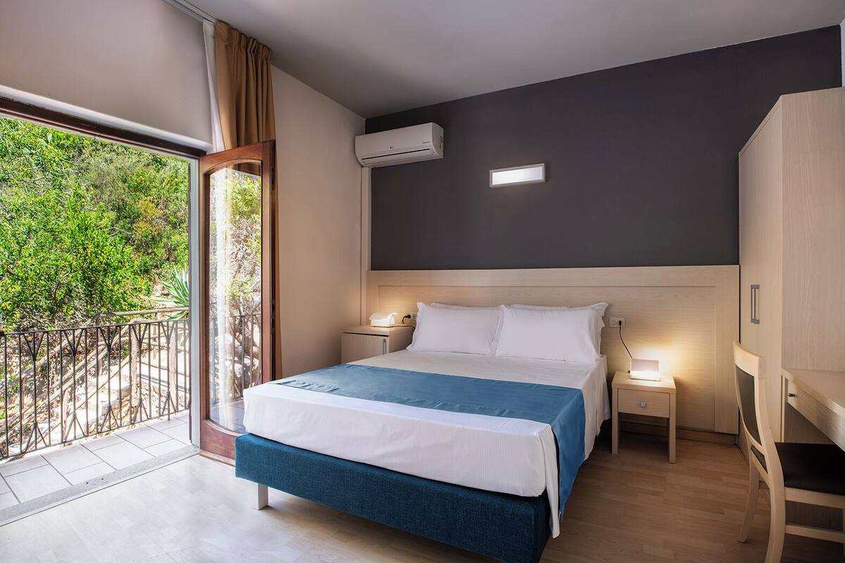 Een hotelkamer met een bed, nachtkastjes, een airconditioning en uitzicht op groen via een balkon.