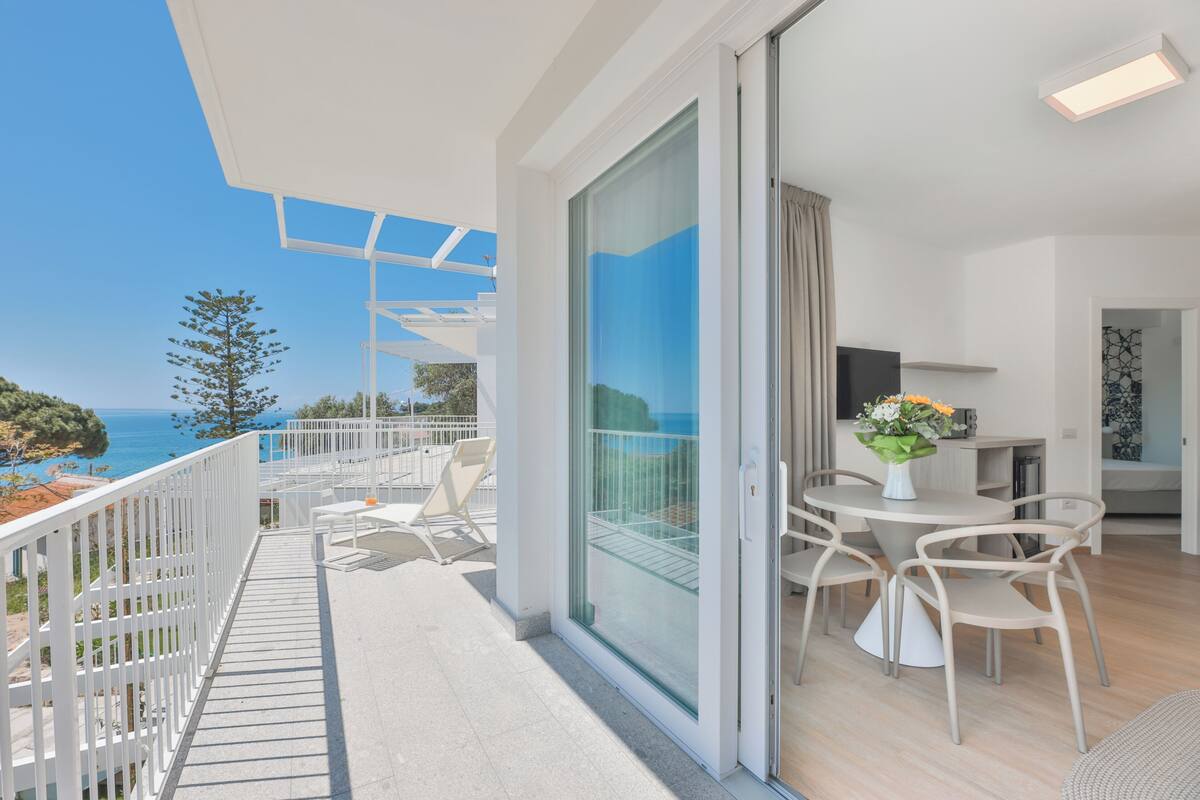 Een balkon met uitzicht op zee, een eethoek met stoelen en een tafel, en een schuifdeur die naar een kamer leidt.