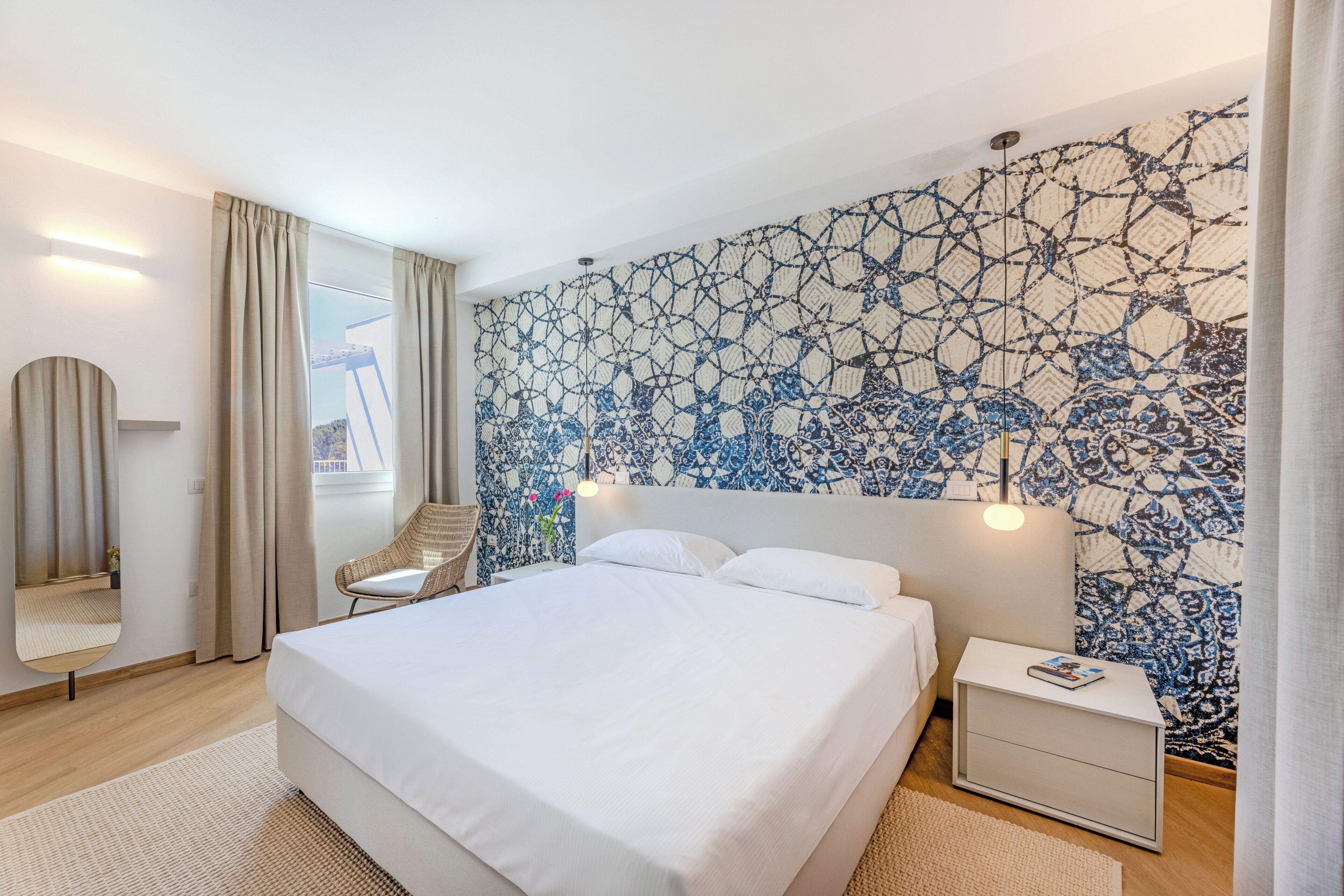 Une chambre d’hôtel moderne avec un grand lit, une table de chevet sur laquelle repose un livre, un mur orné d’un motif bleu et blanc, et une fenêtre avec des rideaux.