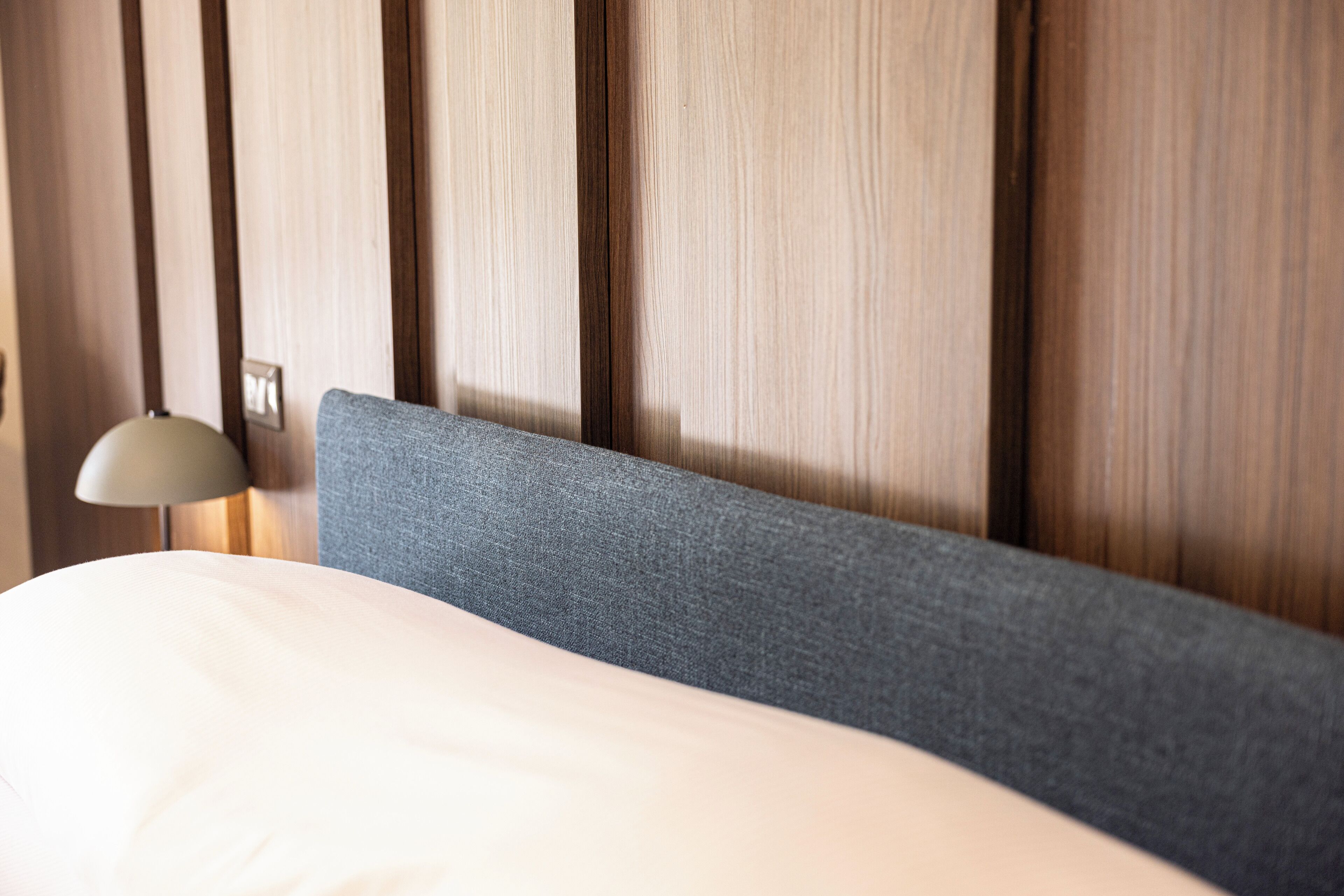 A bed with a grey headboard and a white pillow against a wooden panel wall.