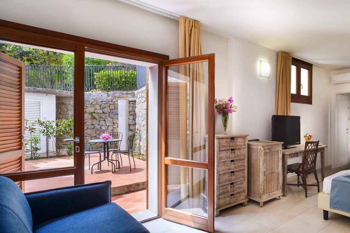 Een slaapkamer met een bed, een bureau met een computer, een televisietoestel en een kledingkast. Er is een balkon met een tafel en stoelen.