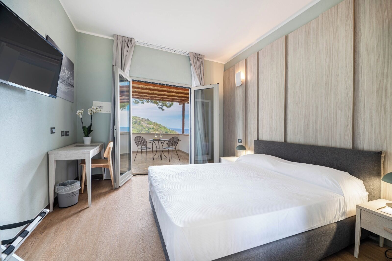 Ein modernes Hotelzimmer mit einem großen Bett, einem Schreibtisch mit Stuhl, einem kleinen Tisch für zwei Personen und einem Balkon mit Blick auf die Hügel.