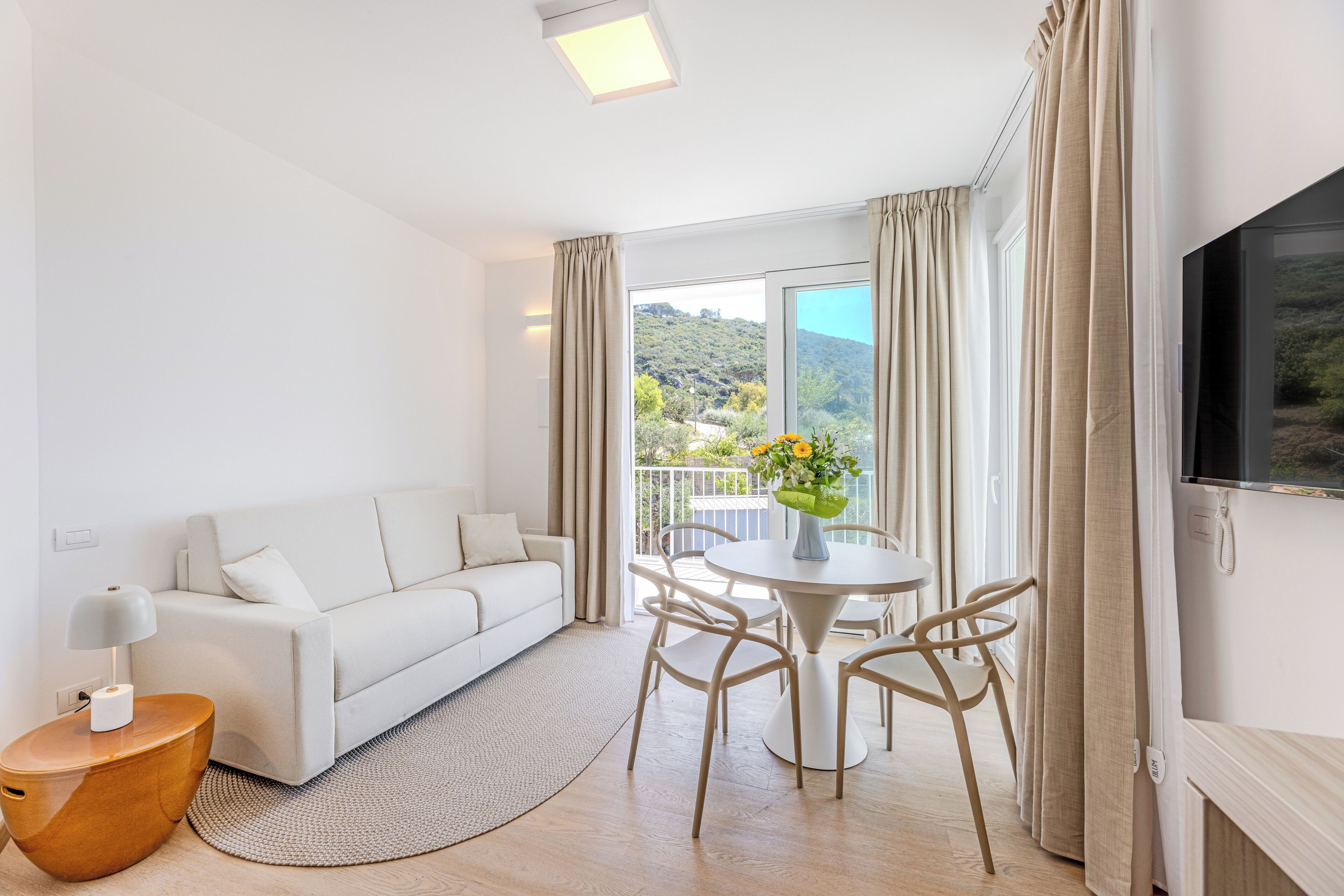 Un salon moderne avec un canapé blanc, une table ronde entourée de chaises, un téléviseur fixé au mur et un balcon agrémenté de rideaux.