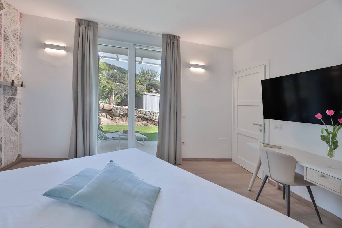 Een moderne hotelkamer met een bed, een bureau, een stoel, een televisie en een glazen schuifdeur naar een buitenruimte.