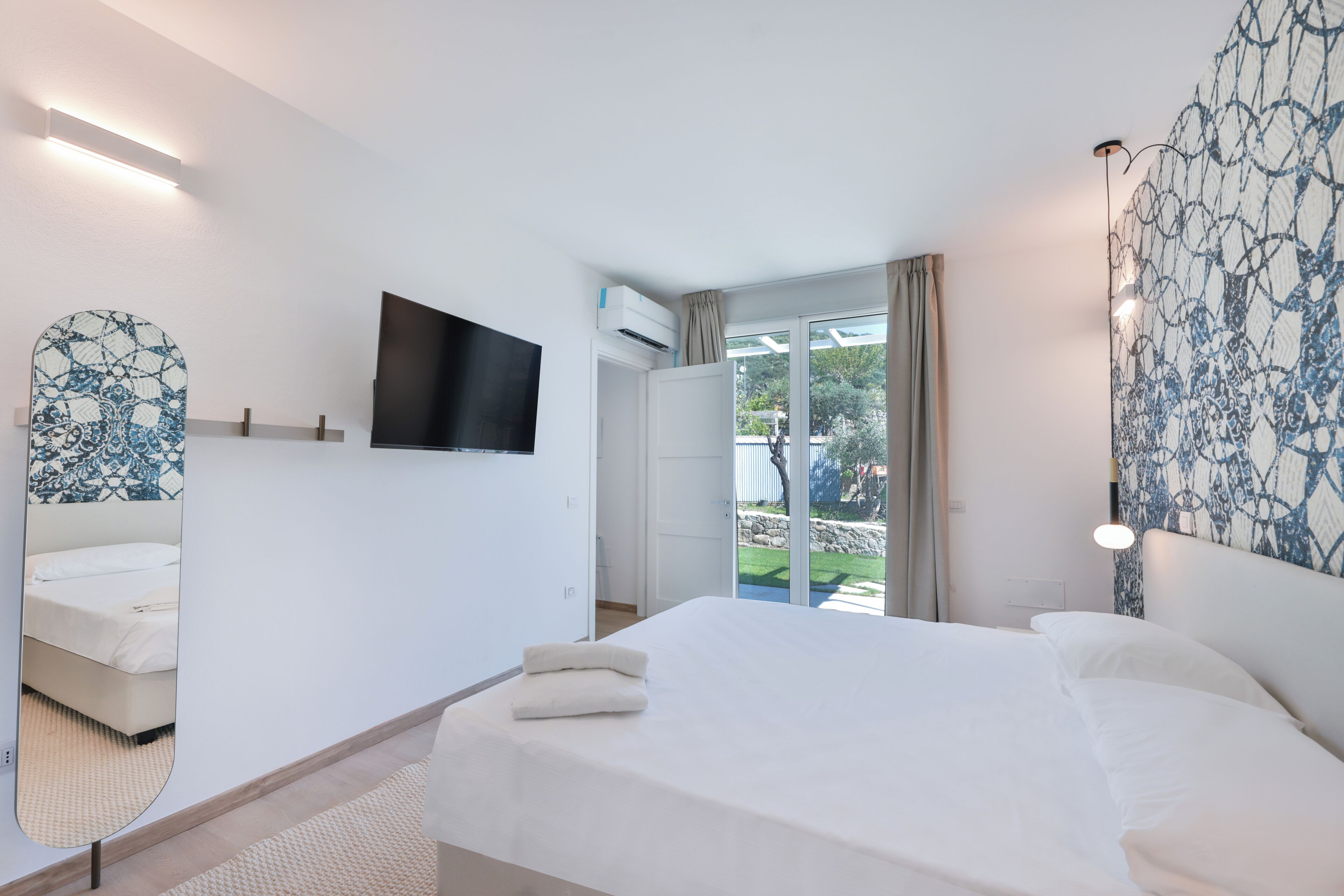 A modern hotel room with a large bed, a flat-screen TV, a large mirror, and a view of the outdoors.