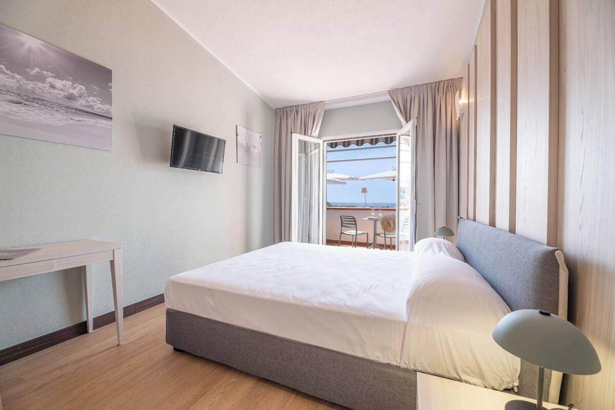 Een hotelkamer met een bed, een bureau, een televisie en een balkon met uitzicht op zee.