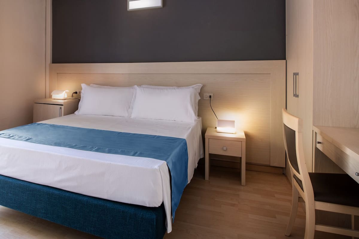 Een keurig opgemaakt bed met een blauw-witte sprei, twee nachtkastjes met lampen en een houten kledingkast.