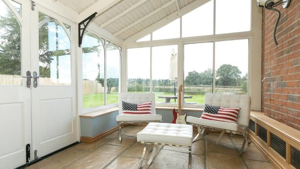A conservatory with a white door, two white cushioned chairs with American flag pillows, a guitar, and a brick wall. - Bournes Farmhouse (Henley-on-Thames)
