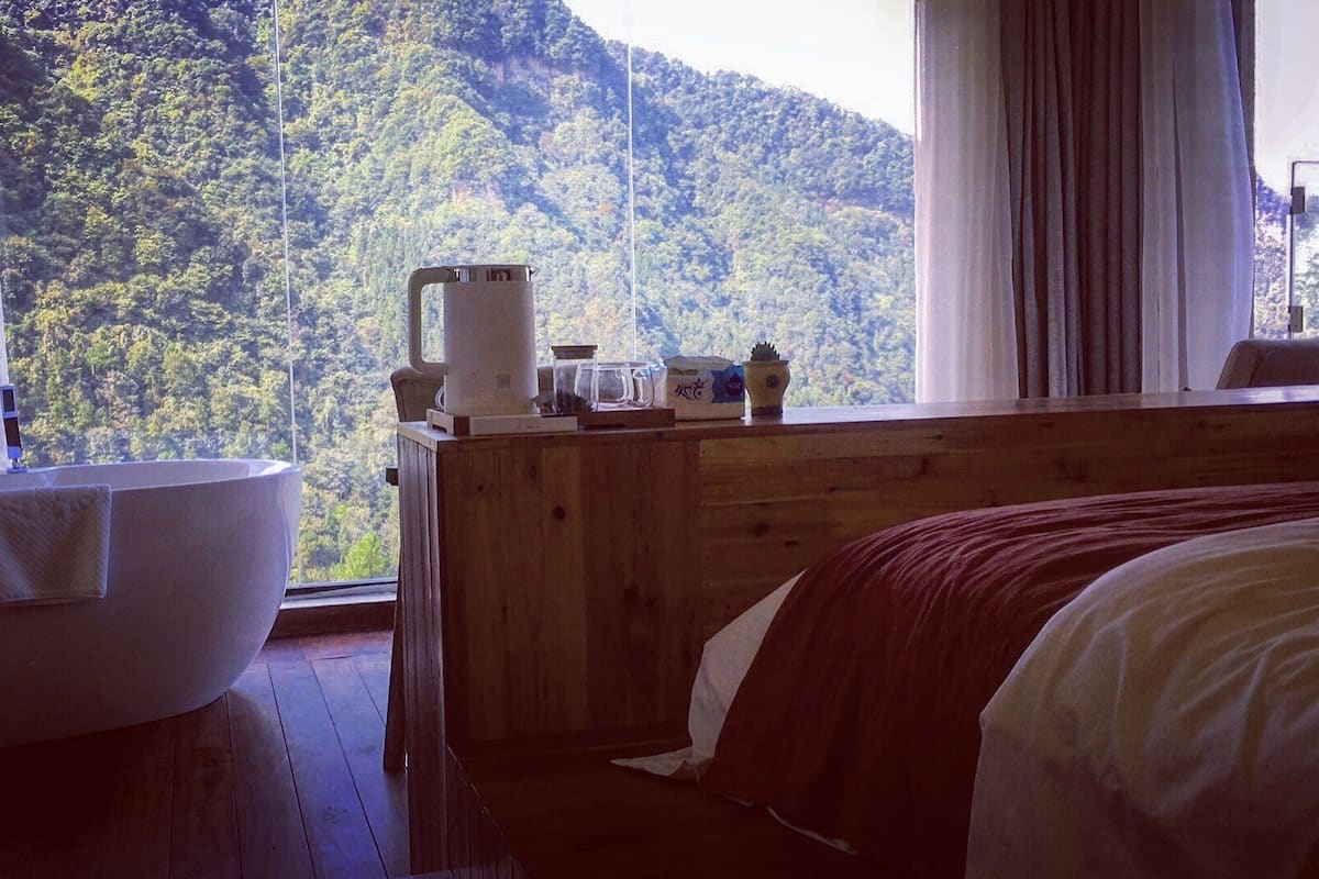 A room with a wooden bed, a large window with a view of a forested mountain, a bathtub, and a wooden dresser with various items on it.