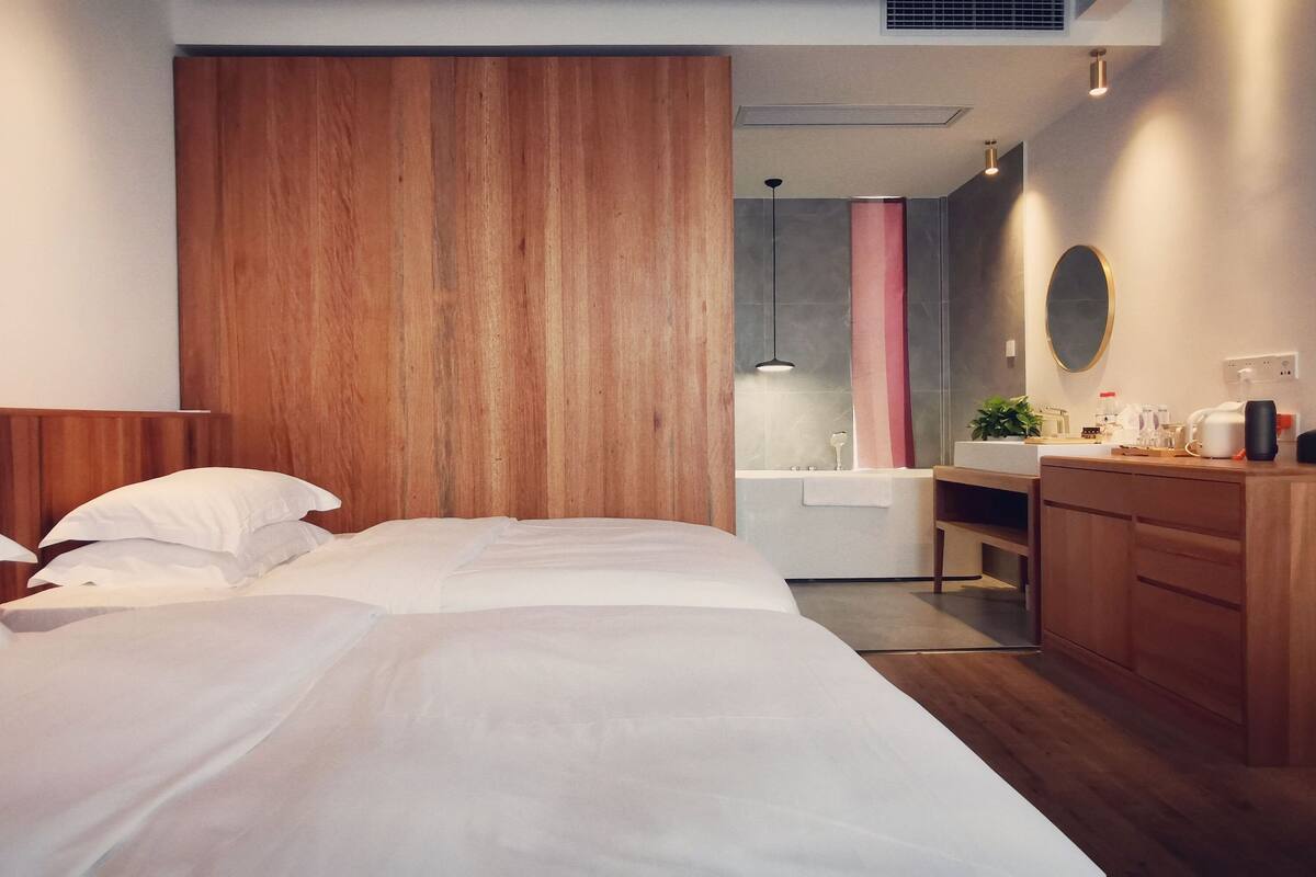 A modern hotel room with two beds, a wooden headboard, a bedside table with a lamp, a small desk, a mirror, and a bathroom with a sink and shower.
