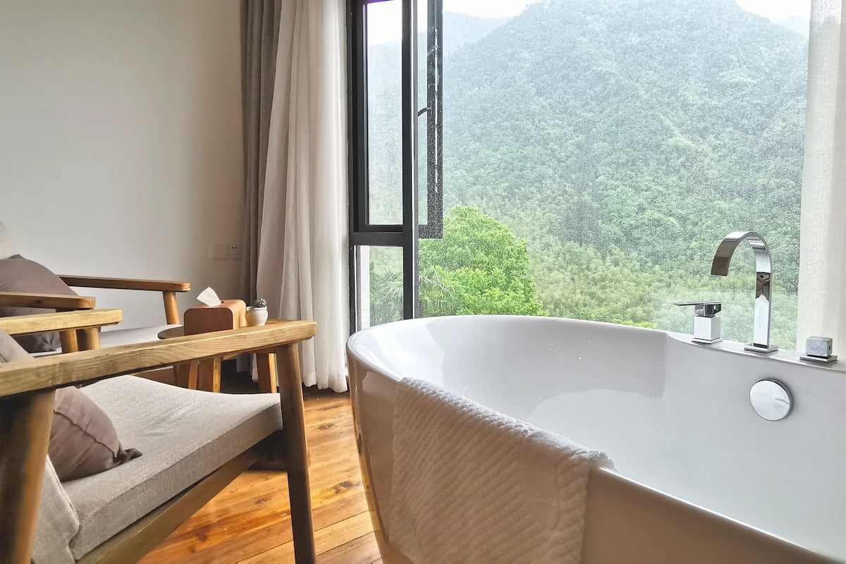 A modern bathroom with a large bathtub, wooden furniture, and a view of a forested mountain.