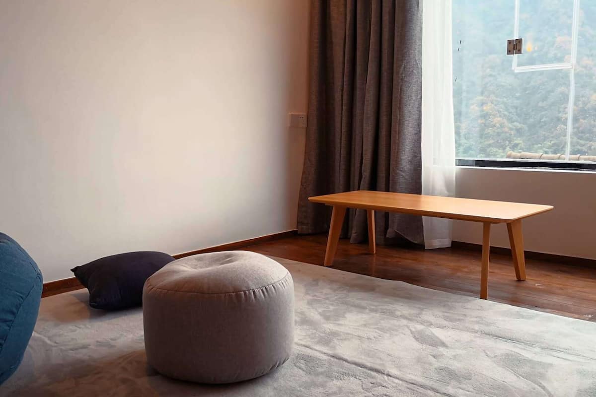 A room with a wooden bench, a round ottoman, a bean bag, and a view of a forest through a window.