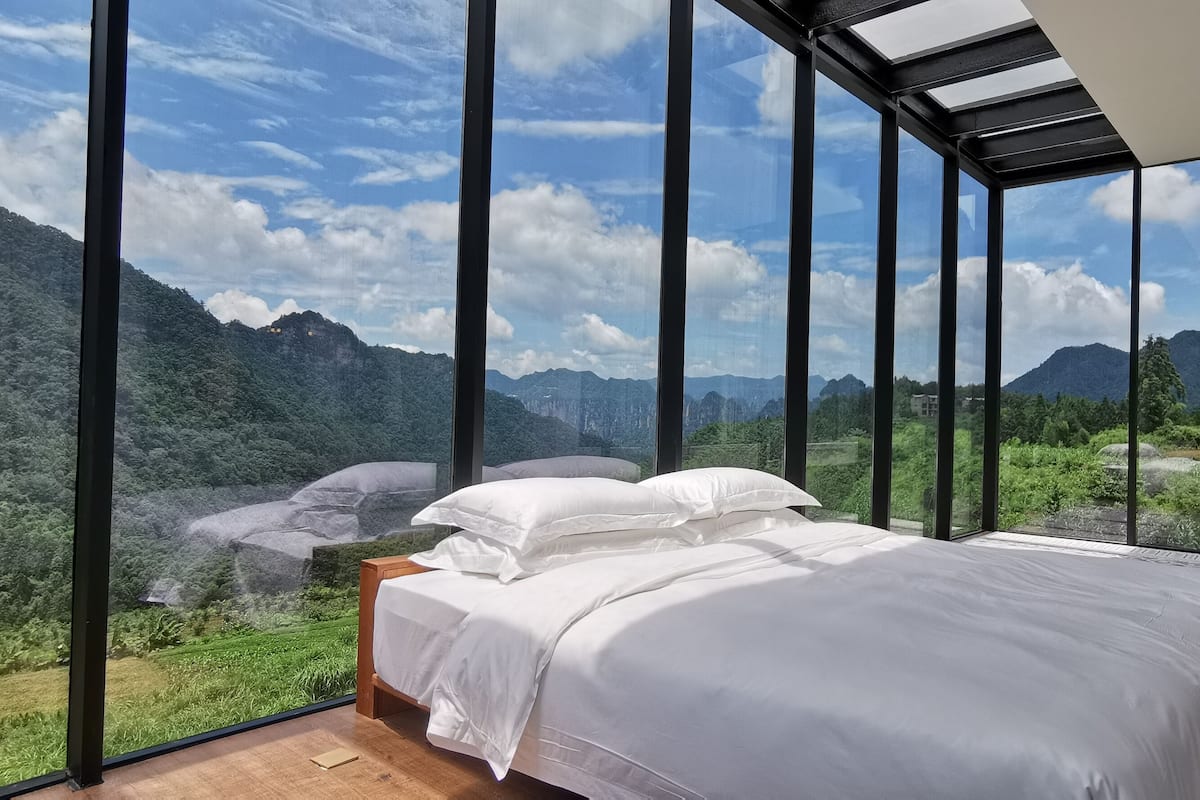A bedroom with a large bed, a wooden nightstand, and a view of a mountainous landscape through floor-to-ceiling windows.