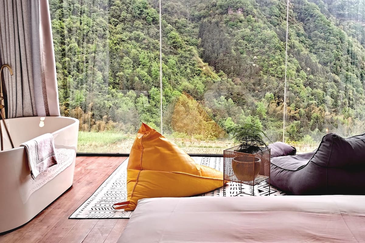 A bedroom with a large window, a bathtub, a bed, a yellow bean bag, a small potted plant, and a wooden cabinet.