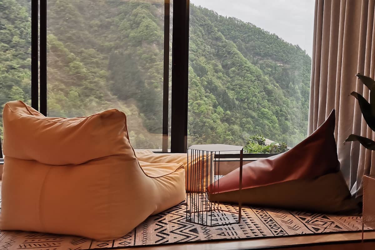 A room with a large window offering a view of a forested mountain. There is a beige sofa, a red bean bag, and a potted plant on a patterned rug.