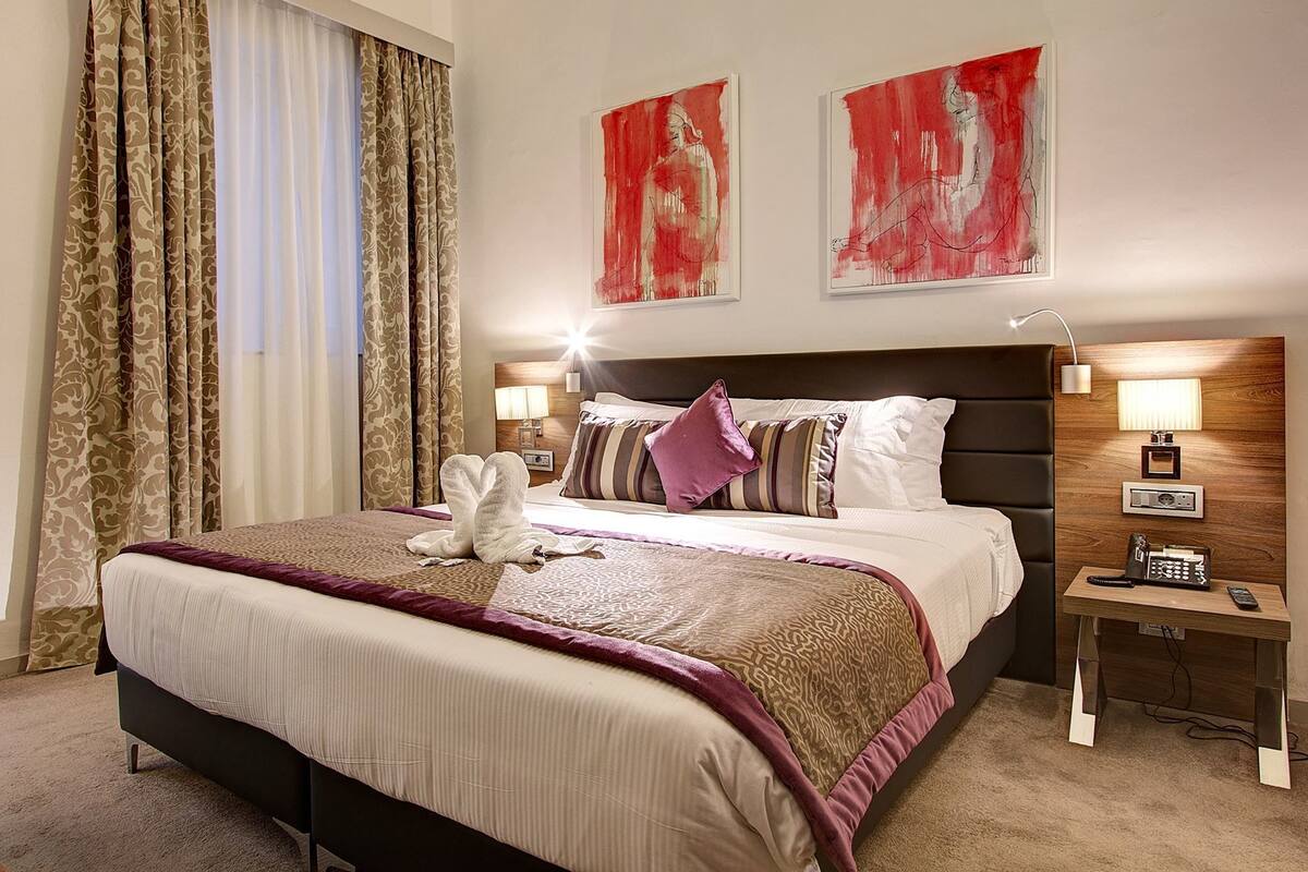 A hotel room with a large bed, two bedside tables, a nightstand, and two abstract paintings on the wall.