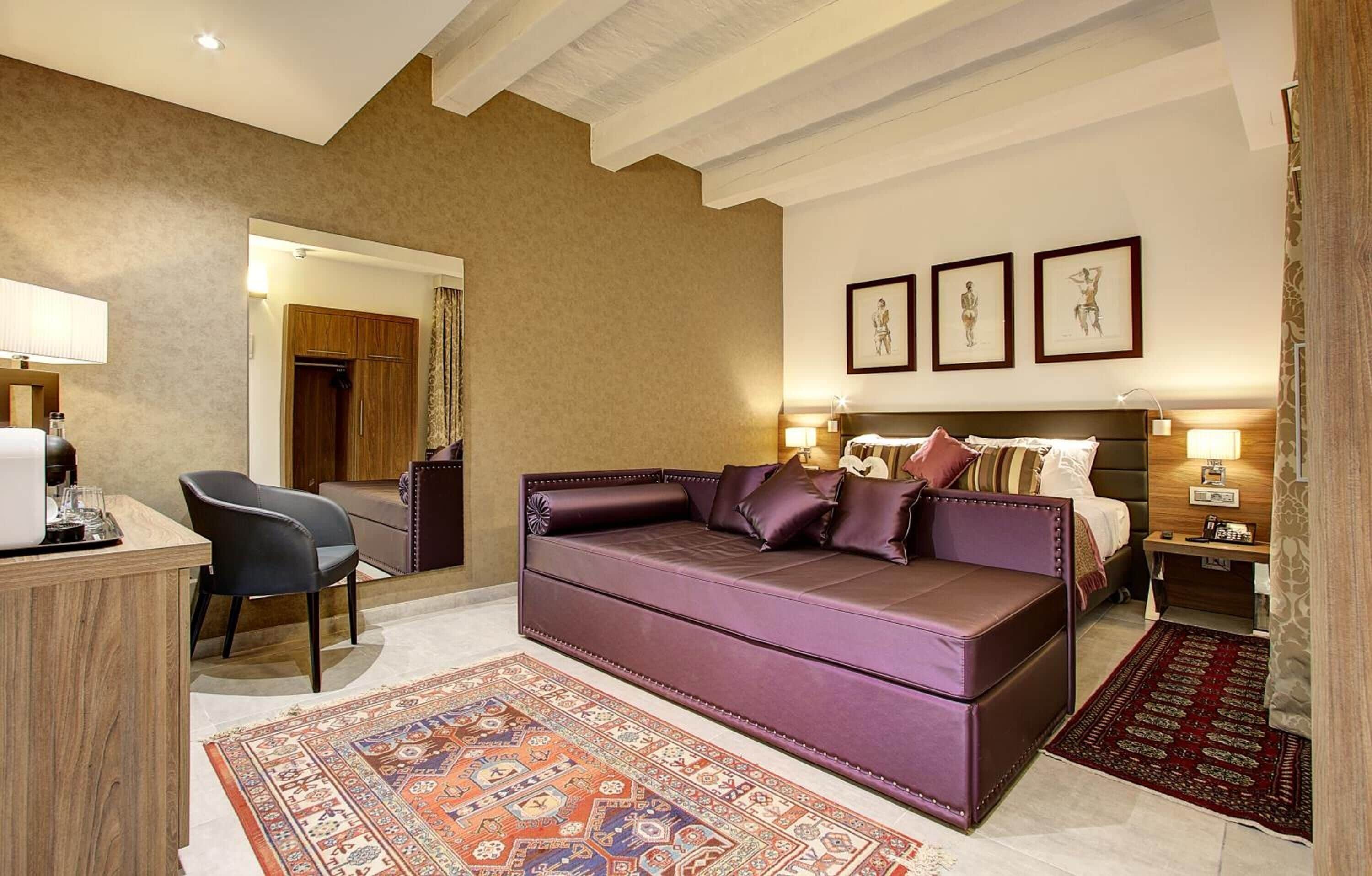 A hotel room with a large purple sofa, a wooden desk, a chair, a bed with a headboard, and a patterned rug.