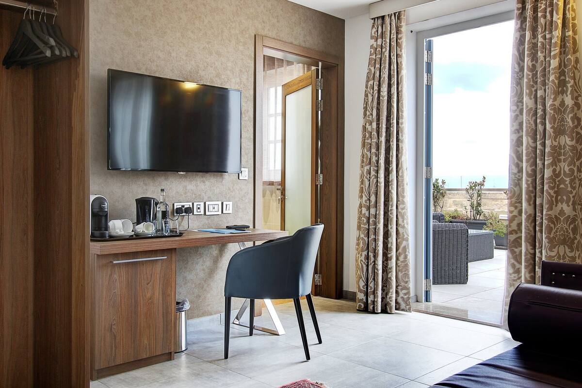 A modern hotel room with a flat-screen TV, a wooden desk with a chair, a balcony with outdoor furniture, and a sliding glass door.