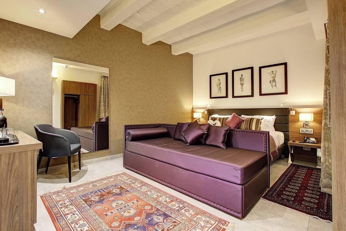 A modern hotel room with a leather sofa, a desk with a coffee machine, a patterned rug, and framed artwork on the wall.