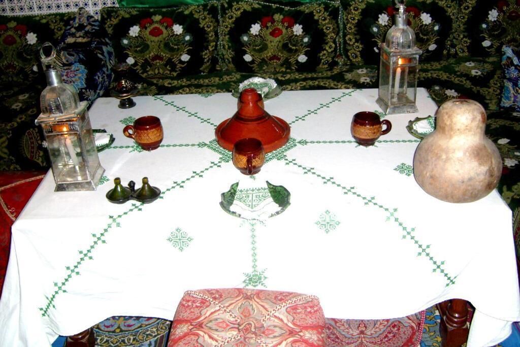 A table with a white tablecloth and green embroidery, surrounded by various items including a red pot, two glasses, and a lantern.