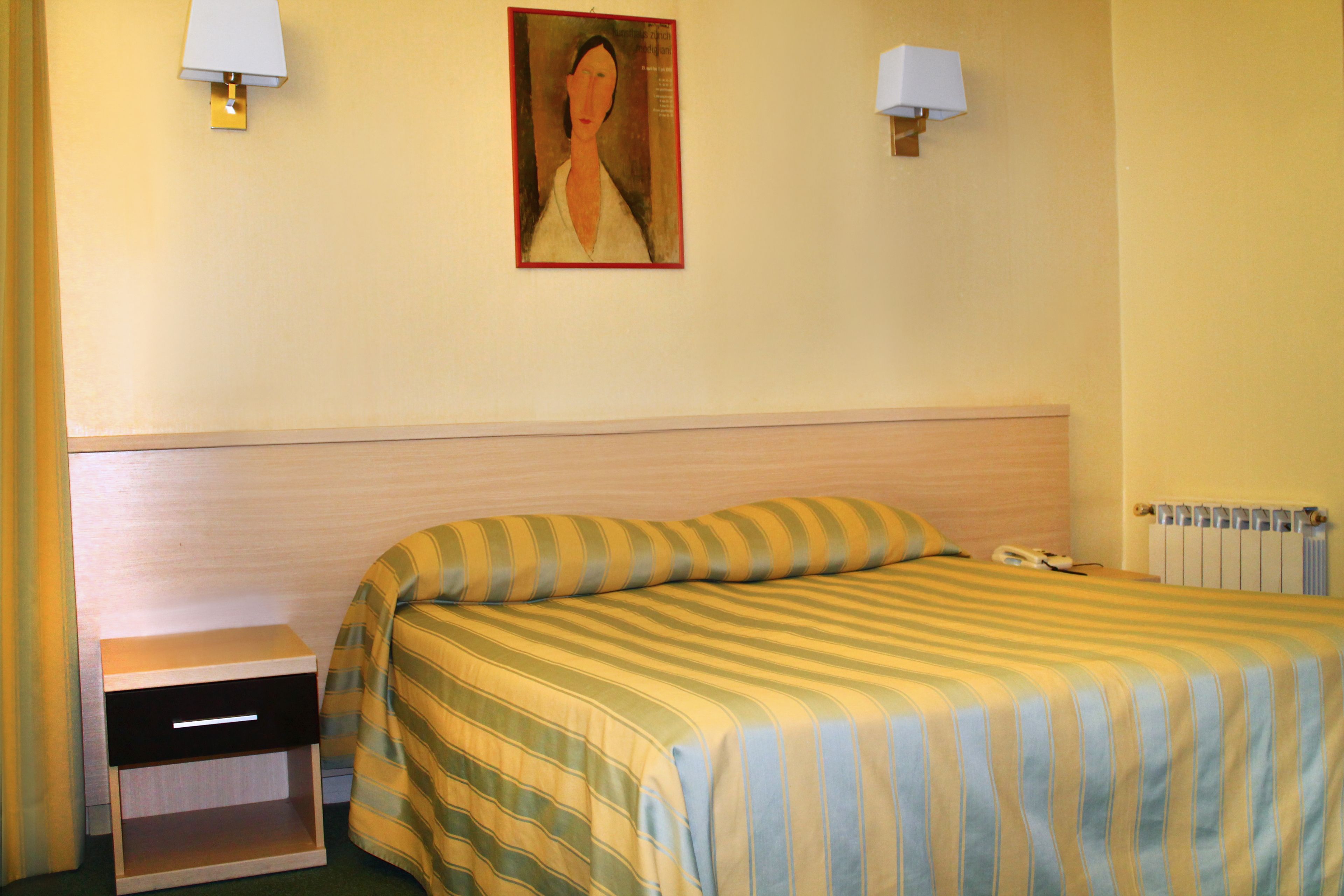 Hotellihuone, jossa on sänky, yöpöytä, lamppu ja taulu seinällä.